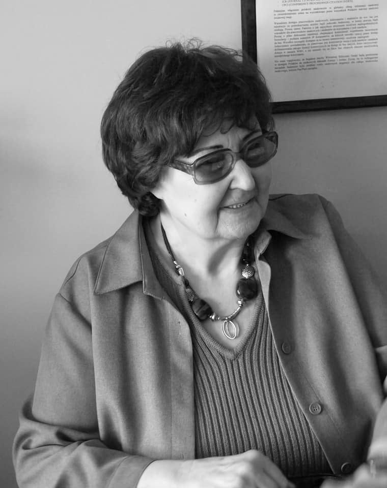 Na czarno-białym zdjęciu prof. Vera Mitrinović patrzy gdzieś w bok przez przyciemnione okulary, na twarzy ma serdeczny uśmiech. Jest ubrana w prążkowaną bluzkę, na którą ma zarzuconą koszulę, a na szyi duże korale.